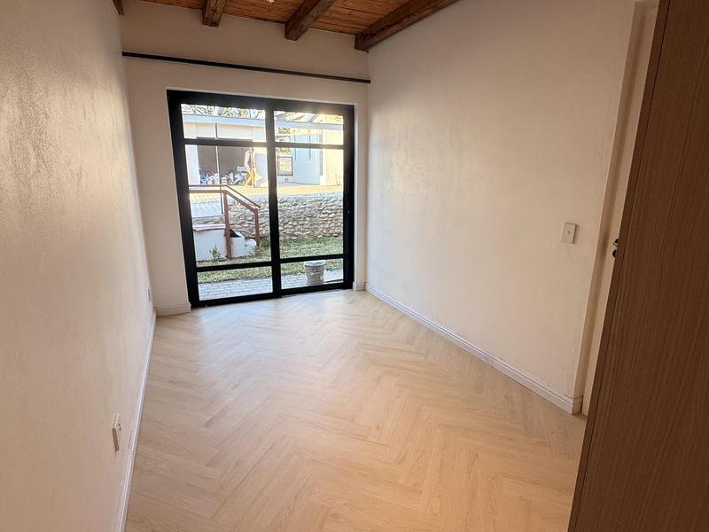 A bright, empty bedroom featuring modern herringbone laminate flooring, exposed timber ceiling beams, and a large black-framed glass sliding door leading to the exterior.