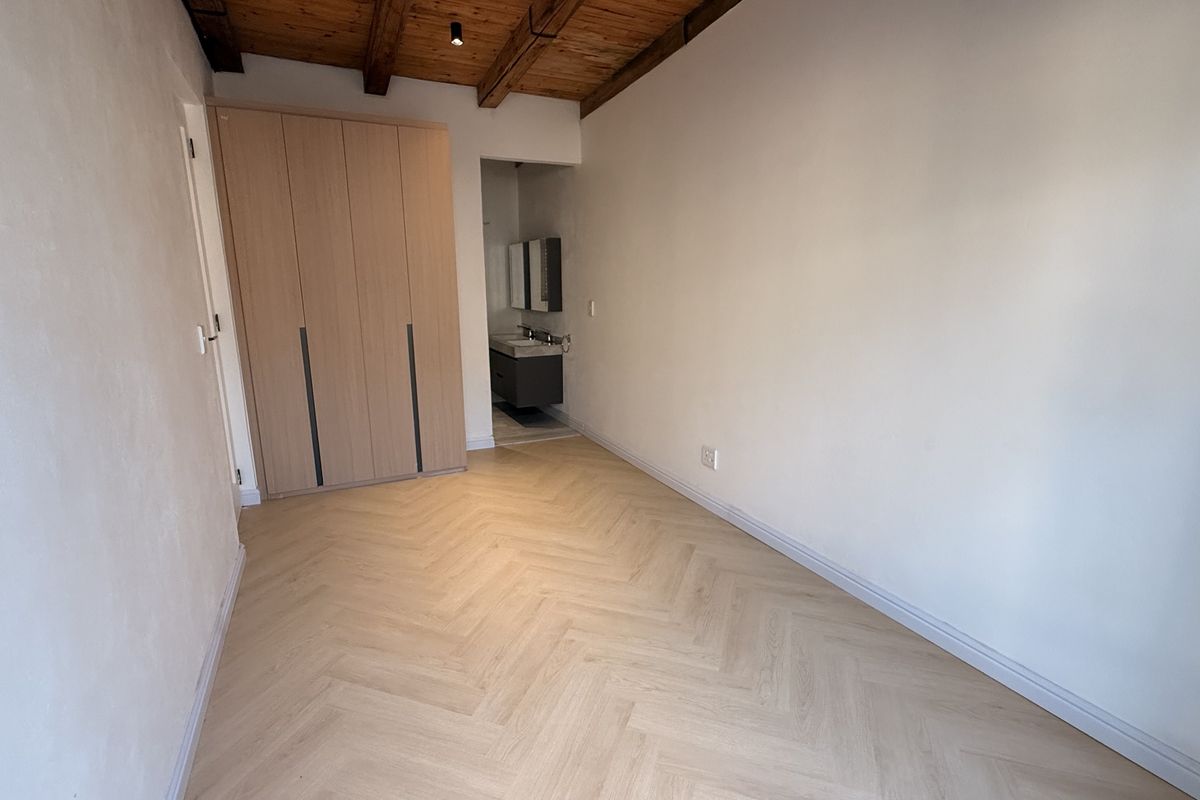 A bright, modern bedroom featuring herringbone flooring, exposed timber ceiling beams, a built-in wardrobe, and an integrated en-suite vanity area.