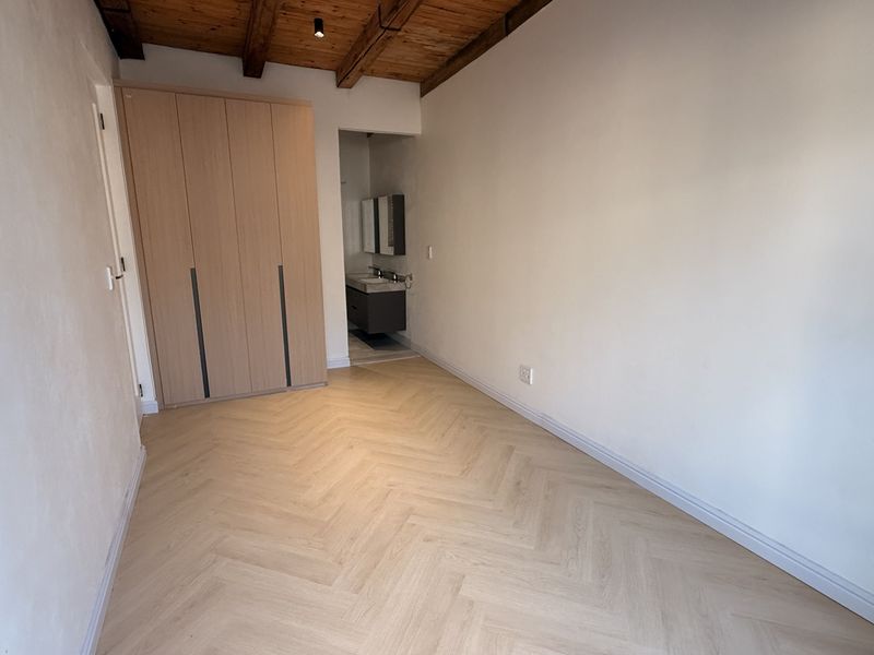 A bright, modern bedroom featuring herringbone flooring, exposed timber ceiling beams, a built-in wardrobe, and an integrated en-suite vanity area.