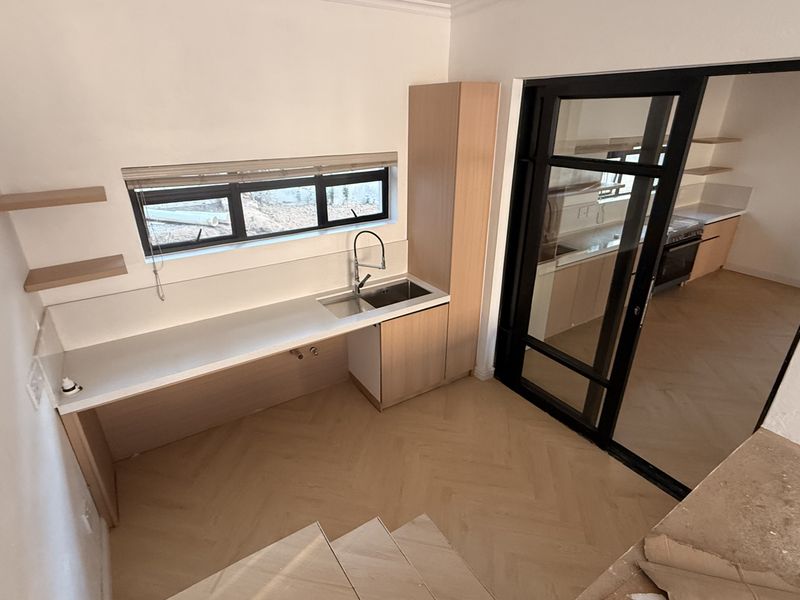 A modern, clean scullery area featuring light timber cabinetry, herringbone flooring, a stainless steel sink, and a black-framed glass door leading to the kitchen.