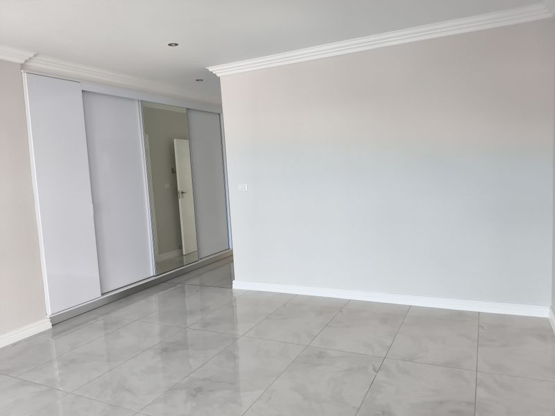 A bright, empty bedroom featuring modern large-format tiled flooring, crown molding, recessed lighting, and a spacious built-in wardrobe with a mirrored sliding door.
