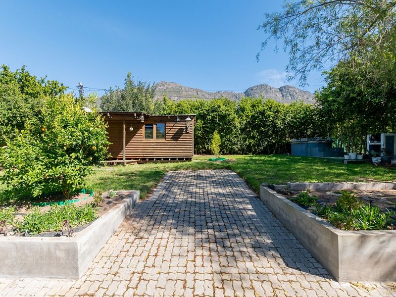 A spacious garden featuring a paved walkway, concrete planter boxes, a timber cabin, and an outdoor braai area set against a mountain backdrop.
