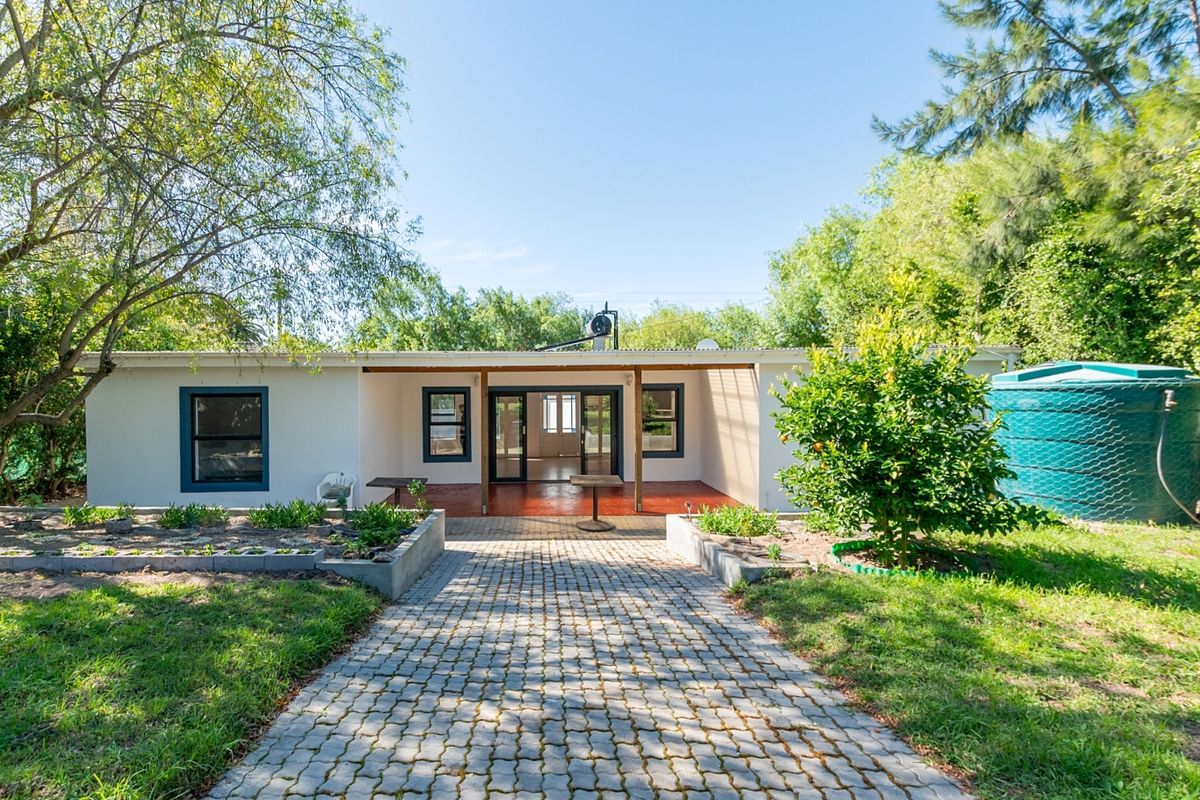Rear view of the house featuring a paved walkway leading to a covered patio, raised garden beds, and a large water storage tank.