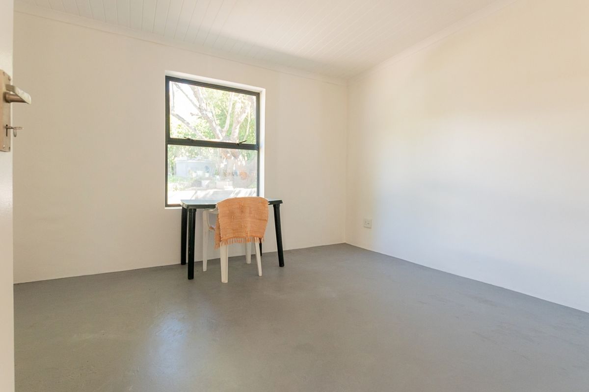 A bright, empty bedroom featuring a painted concrete floor, tongue and groove ceiling, and a large aluminium-framed window.