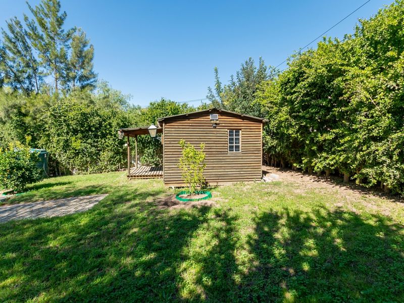 A detached timber cabin with a small covered deck, situated in a private garden setting with mature hedging and paved access.