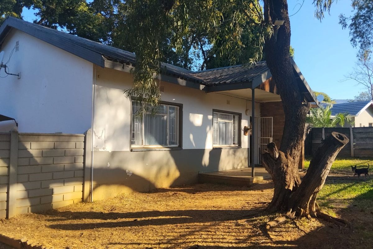 Rear view of a single-story house featuring a covered porch, concrete block boundary wall, and mature trees in the garden area.