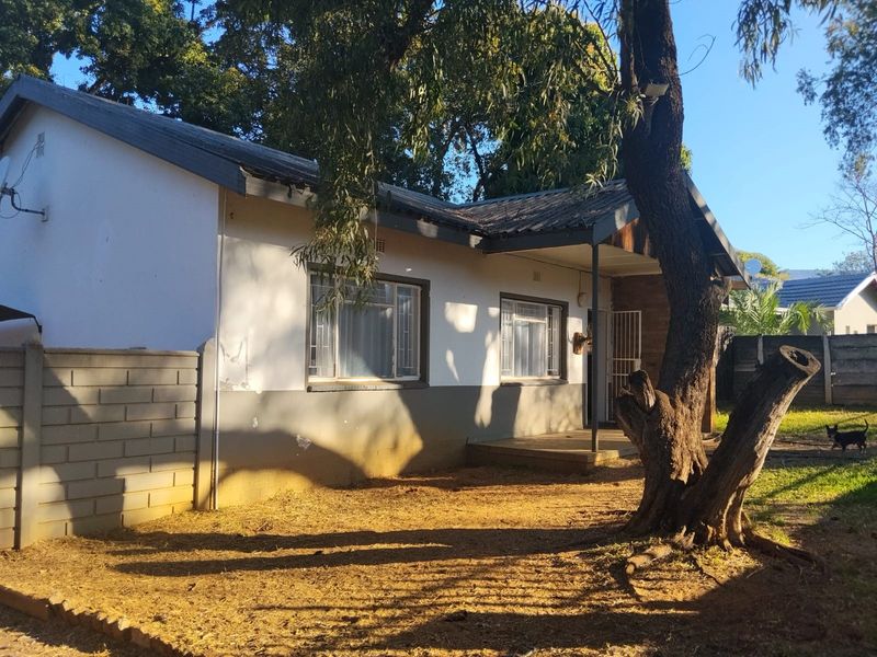 Rear view of a single-story house featuring a covered porch, concrete block boundary wall, and mature trees in the garden area.
