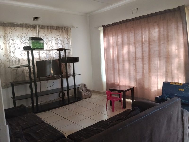 A living room featuring tiled flooring, neutral walls, and large windows with curtain treatments, providing a functional and well-lit living space.