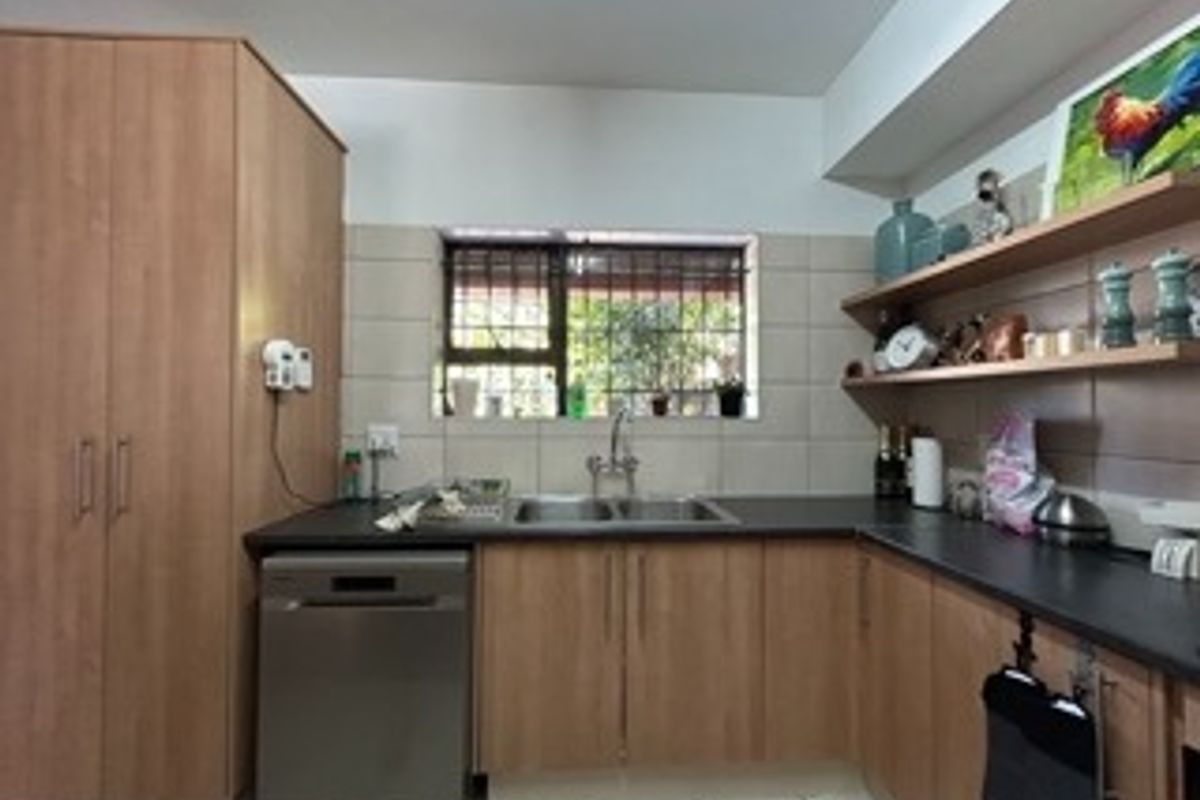 A functional kitchen featuring light wood-look cabinetry, dark countertops, a double basin sink, and open shelving with a tiled backsplash.