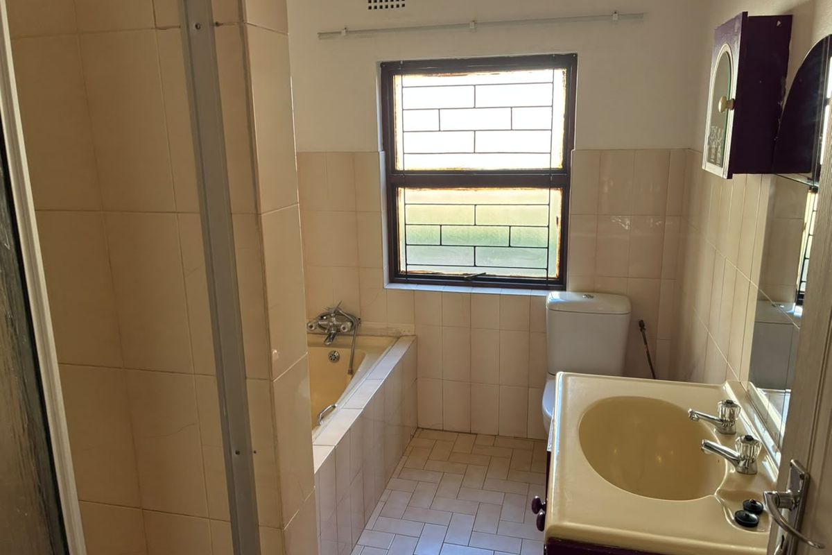 A dated bathroom featuring a built-in bathtub, pedestal basin, and toilet, with neutral wall tiling and security bars on the window.