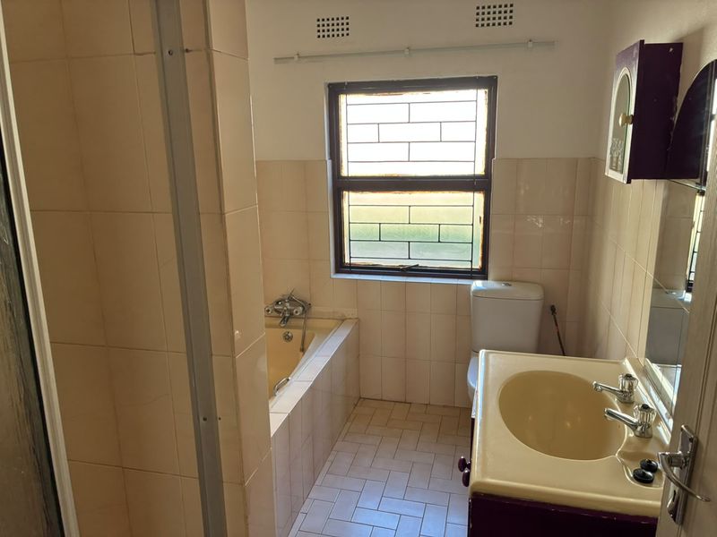 A dated bathroom featuring a built-in bathtub, pedestal basin, and toilet, with neutral wall tiling and security bars on the window.