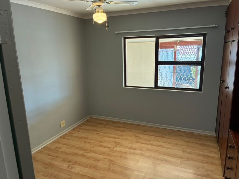 A clean, empty bedroom featuring laminate flooring, built-in cupboards, a ceiling fan, and a window fitted with a security gate.