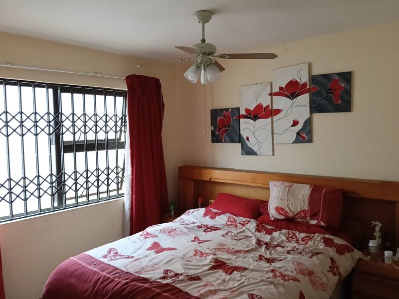 A bedroom featuring a built-in wooden headboard, a ceiling fan, and a window fitted with a security gate.