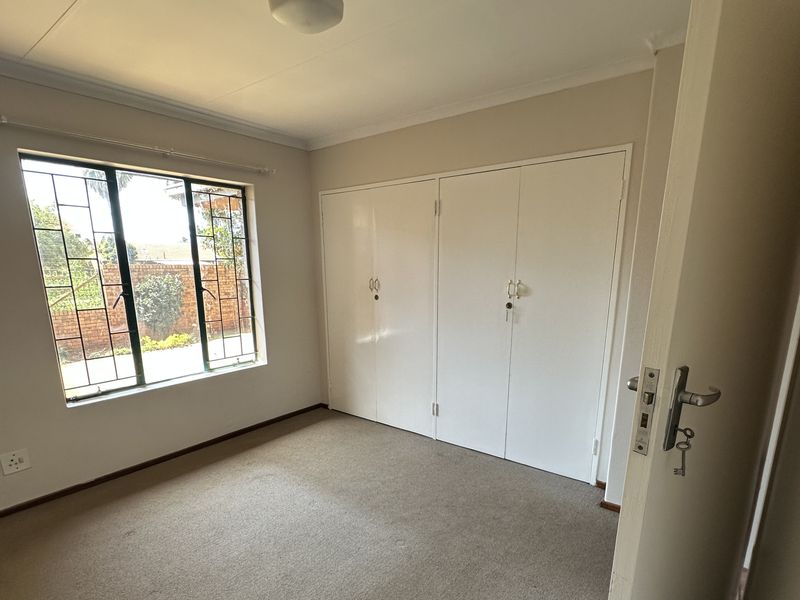 A bright, empty bedroom featuring neutral walls, carpeted flooring, and ample built-in cupboard storage with security bars on the window.