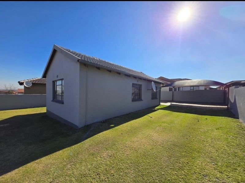 A view of the rear exterior of the house, featuring a well-maintained lawn, perimeter wall, and a covered carport area.