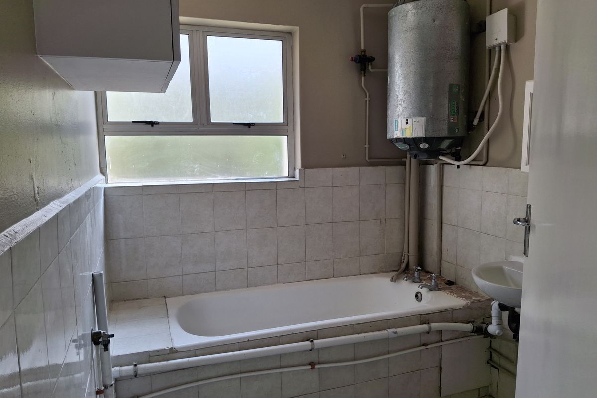 A functional bathroom featuring a built-in bathtub, wall-mounted basin, and exposed geyser unit with tiled splashbacks and a frosted window.