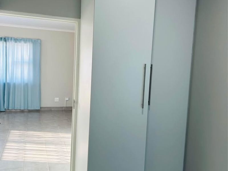 A clean, neutral hallway featuring a tall built-in cupboard with modern handles and light-colored tiled flooring, leading into an adjacent living space.