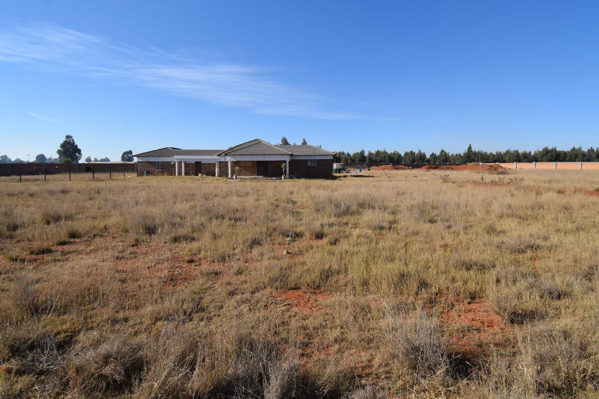 A wide-angle view of a residential property featuring a single-story dwelling set on a large, undeveloped plot of land with perimeter fencing.