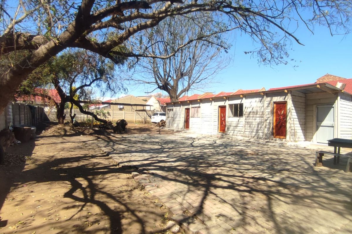 Rear exterior view featuring a paved courtyard area, concrete block outbuildings with timber doors, and mature trees within a fenced property boundary.