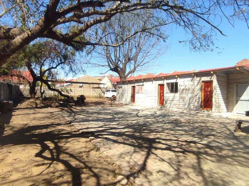 Rear exterior view featuring a paved courtyard area, concrete block outbuildings with timber doors, and mature trees within a fenced property boundary.