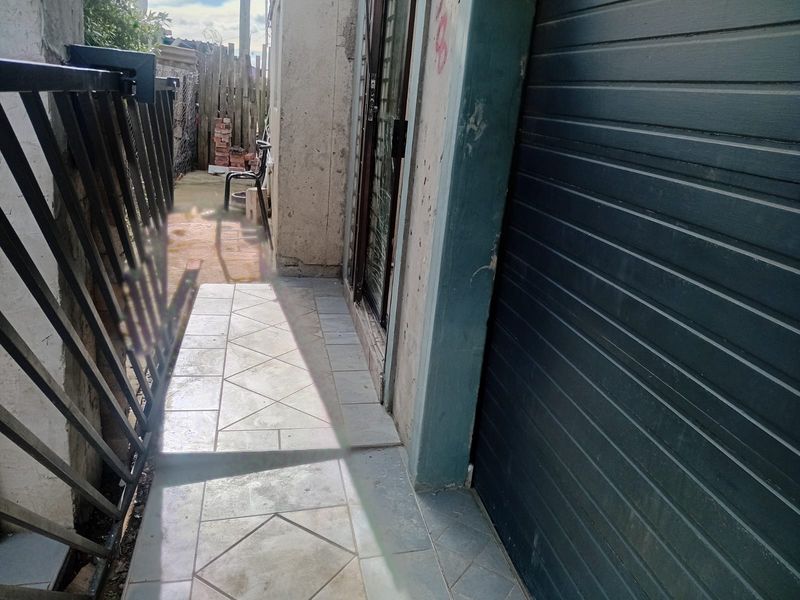 A narrow exterior rear walkway featuring tiled flooring, a metal security gate, and a roller shutter door leading to the property.