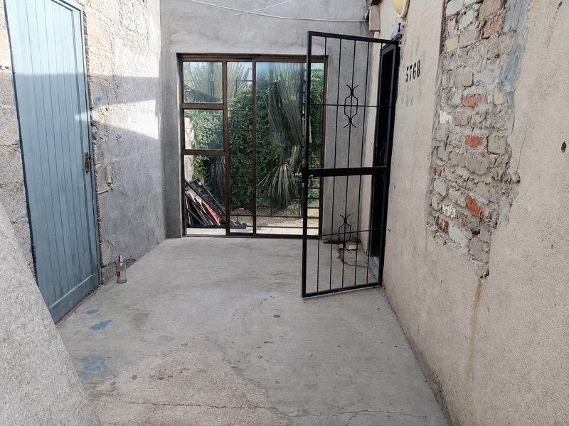 An unfinished outdoor patio area featuring concrete flooring, exposed brick walls, and a security gate leading to a sliding glass door entrance.