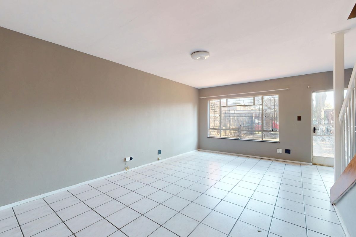 An empty, open-plan living area featuring neutral tiled flooring, a staircase, and natural light through a large window with security bars.