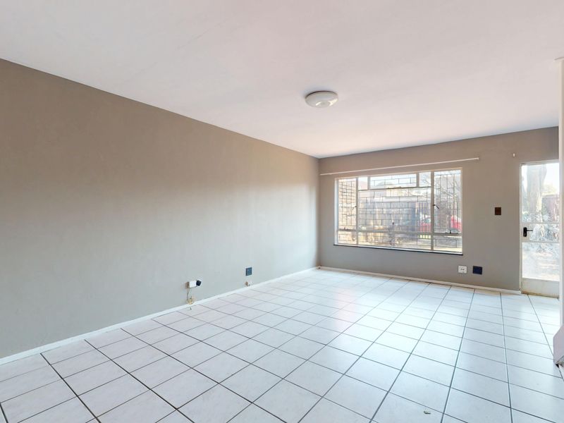 An empty, open-plan living area featuring neutral tiled flooring, a staircase, and natural light through a large window with security bars.