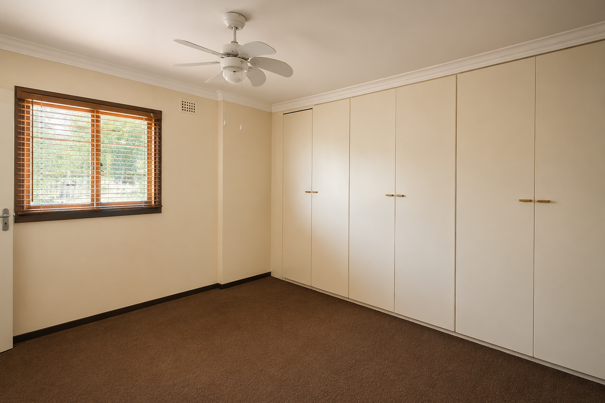 A clean, empty bedroom featuring neutral walls, brown carpet, a ceiling fan, and extensive built-in wardrobe storage.