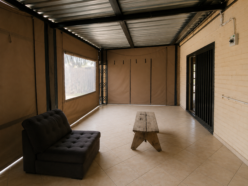 A covered patio area featuring tiled flooring, exposed steel roof structure, and roll-up canvas blinds, providing a versatile outdoor living space.