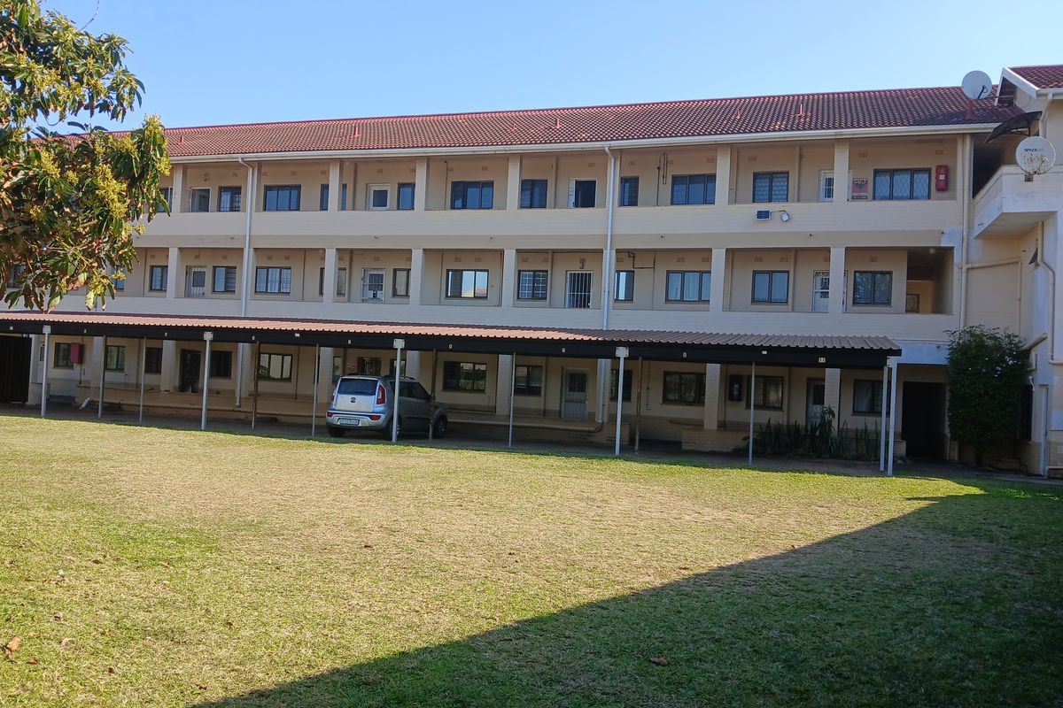 Rear view of a multi-story apartment complex featuring a large lawn area and covered parking bays for residents.
