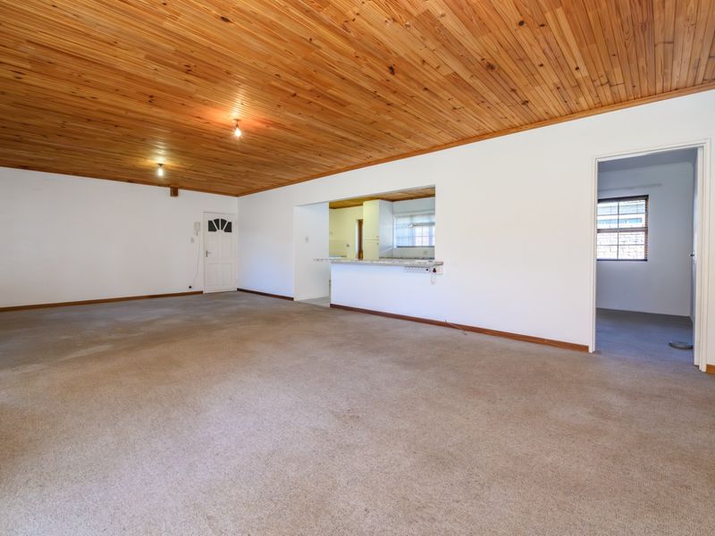 Spacious, empty living area featuring a timber tongue and groove ceiling, carpeted flooring, and a convenient pass-through window connecting to the kitchen.