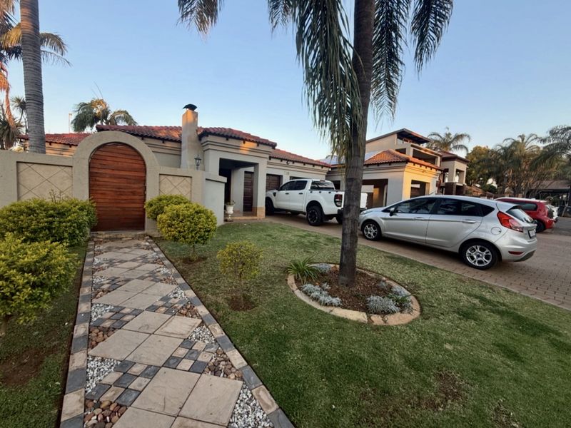 Front elevation of a well-maintained house featuring a paved driveway, stone-tiled walkway, arched wooden gate, and established garden landscaping.