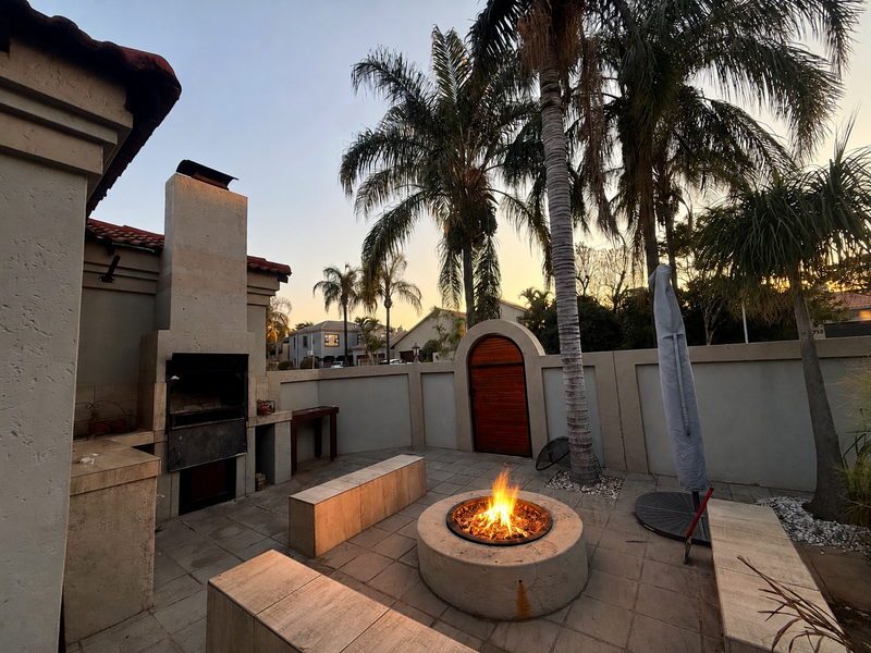 Outdoor patio area featuring a built-in braai, stone fire pit with integrated bench seating, and a secure arched gate within a walled garden setting.