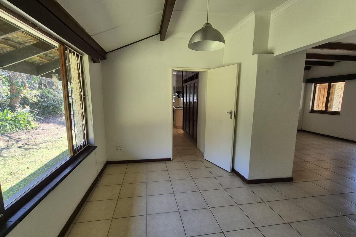 An empty living area featuring tiled floors, exposed timber ceiling beams, and large windows providing natural light and garden views.