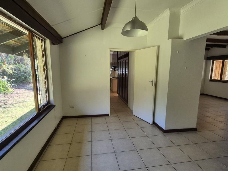 An empty living area featuring tiled floors, exposed timber ceiling beams, and large windows providing natural light and garden views.