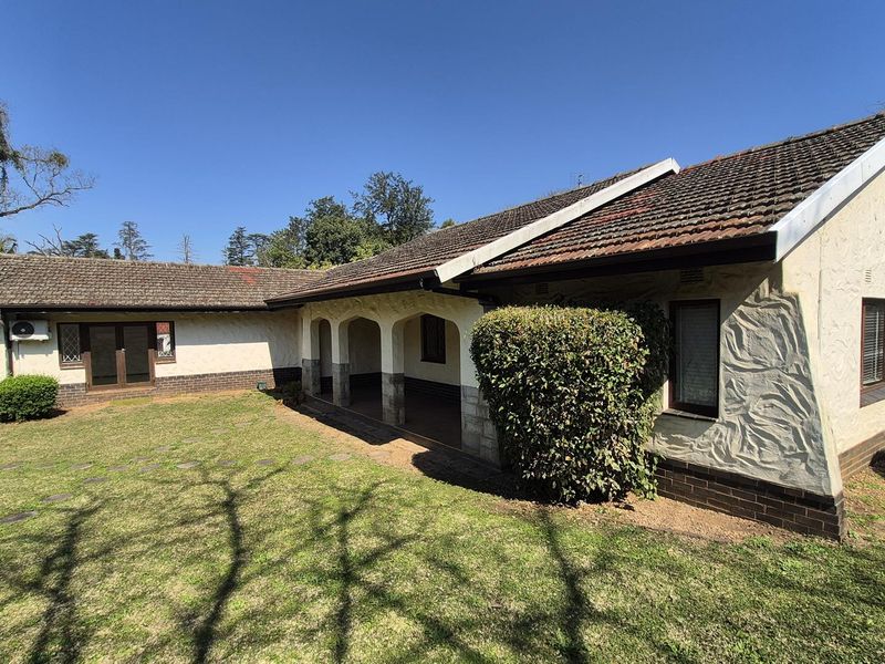 Rear view of a single-story house featuring a covered patio, textured exterior walls, and a tiled roof set within a spacious garden.