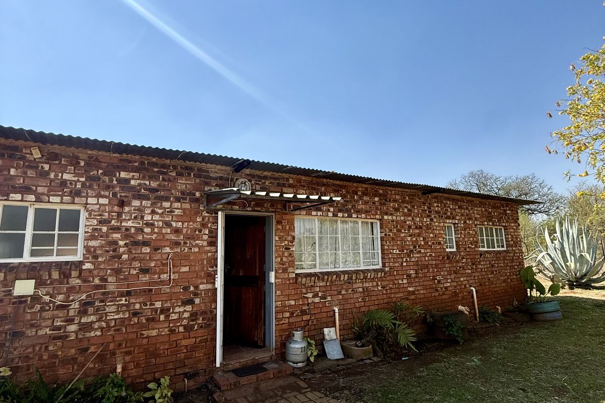 Exterior view of a single-story apartment featuring exposed brick walls, corrugated metal roofing, and grid-pattern windows set within a garden area.