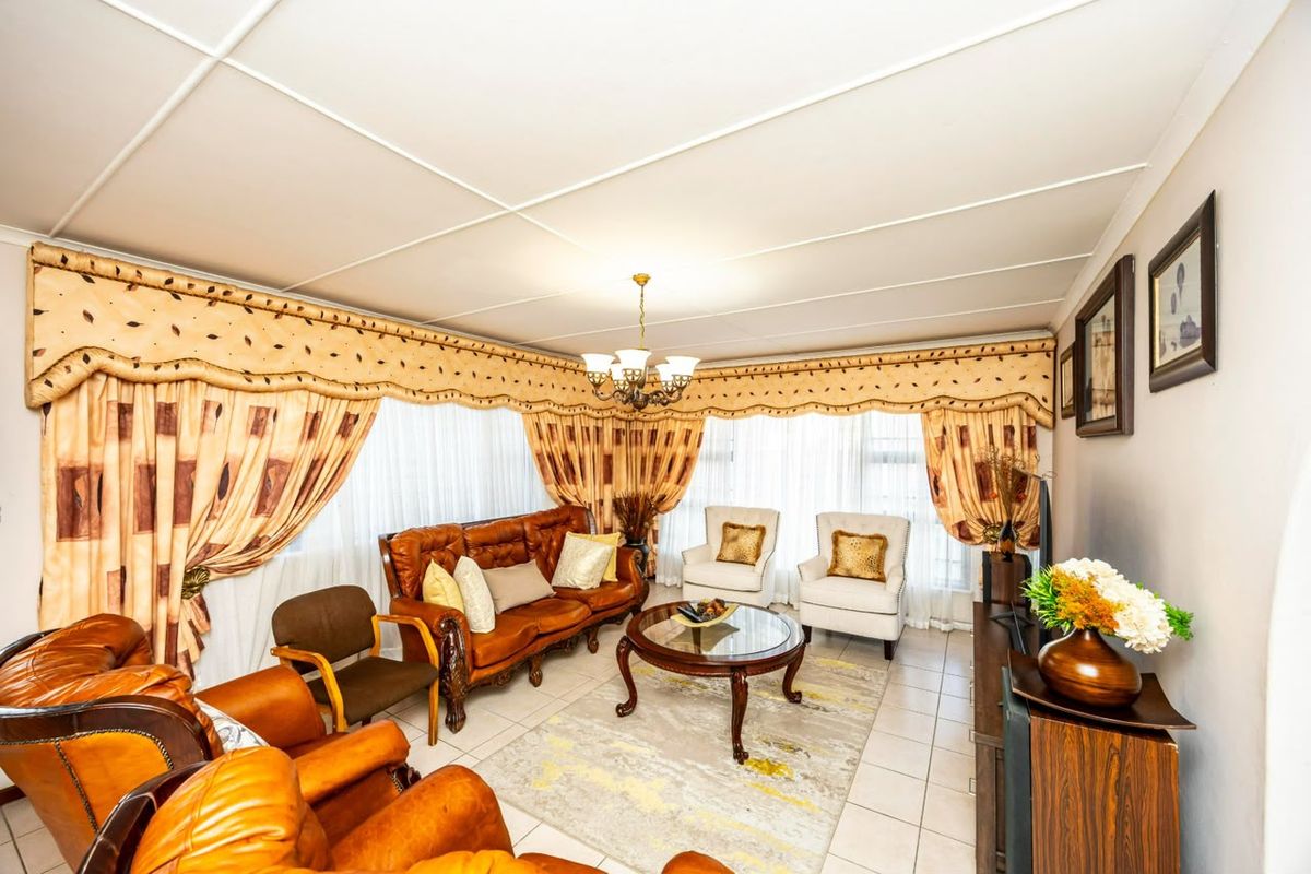 A spacious living room featuring tiled flooring, a central chandelier, and large windows with decorative pelmets.
