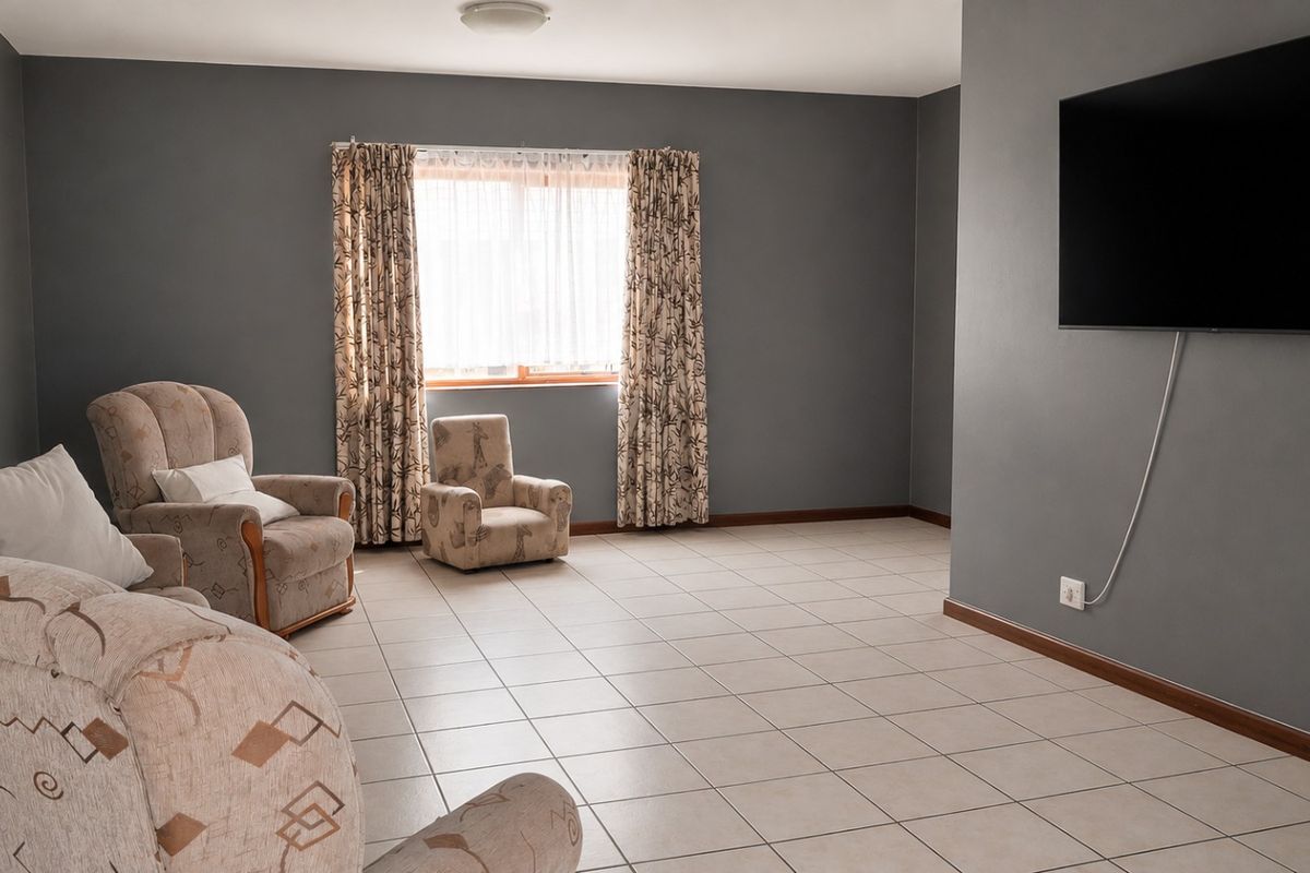 A spacious living room featuring neutral tiled flooring, grey painted walls, and a wall-mounted television setup with natural light from the window.