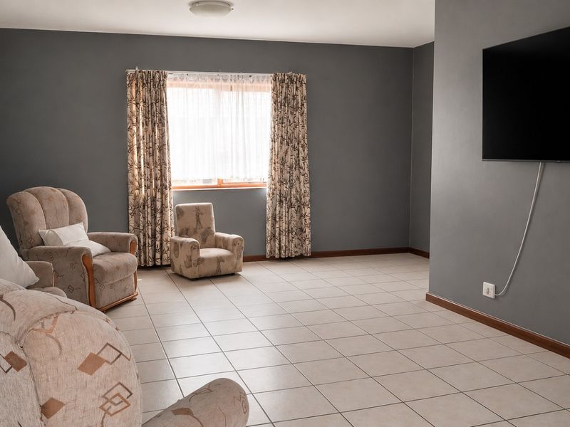 A spacious living room featuring neutral tiled flooring, grey painted walls, and a wall-mounted television setup with natural light from the window.