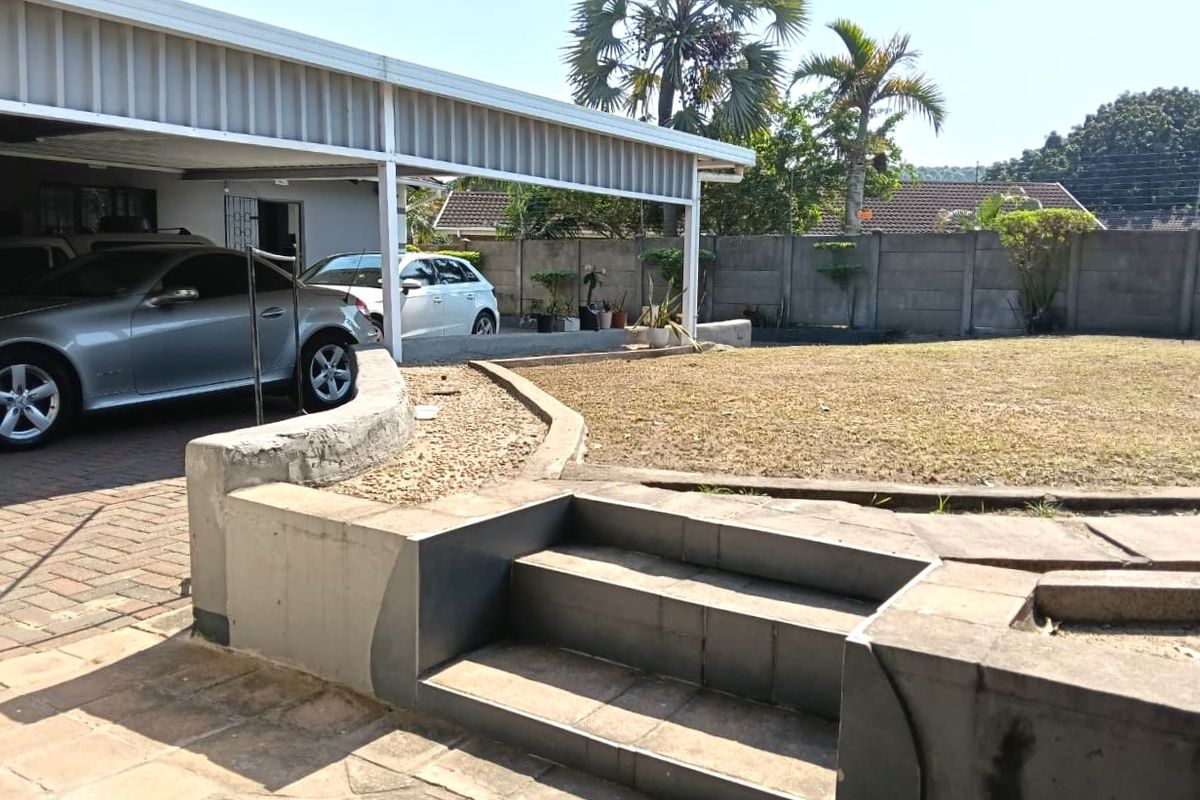 Rear exterior view of the cottage featuring a covered carport, paved driveway, concrete retaining walls with steps, and a fenced garden area.