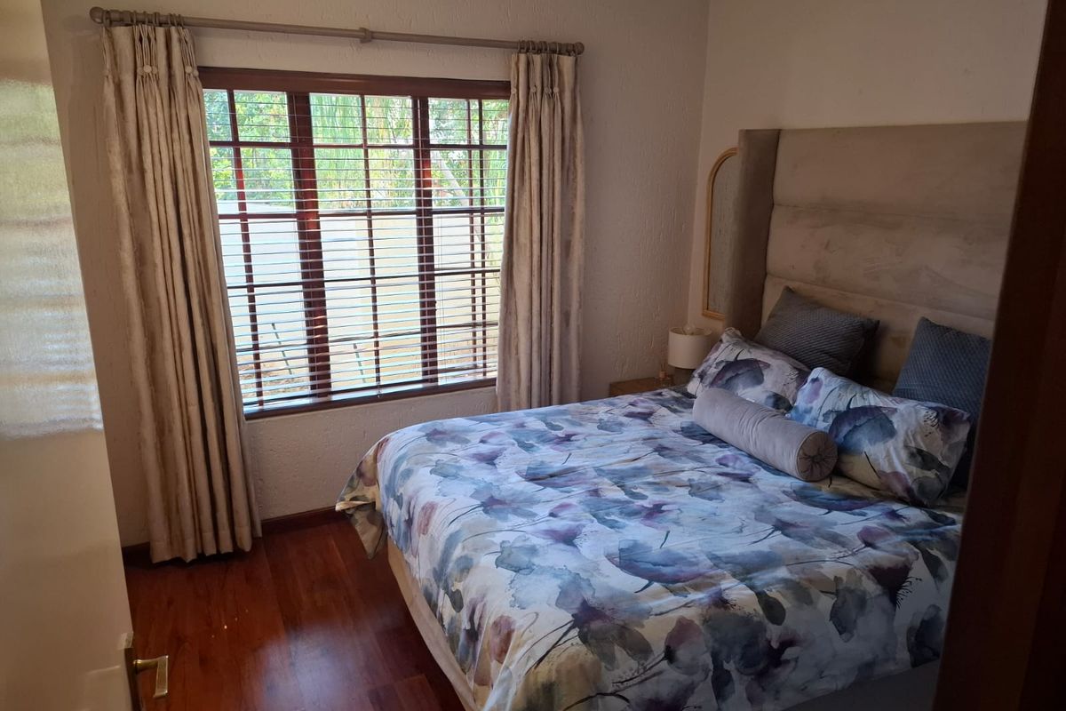 A cozy bedroom featuring warm-toned laminated flooring, a large wooden-framed window with venetian blinds, and neutral wall finishes.