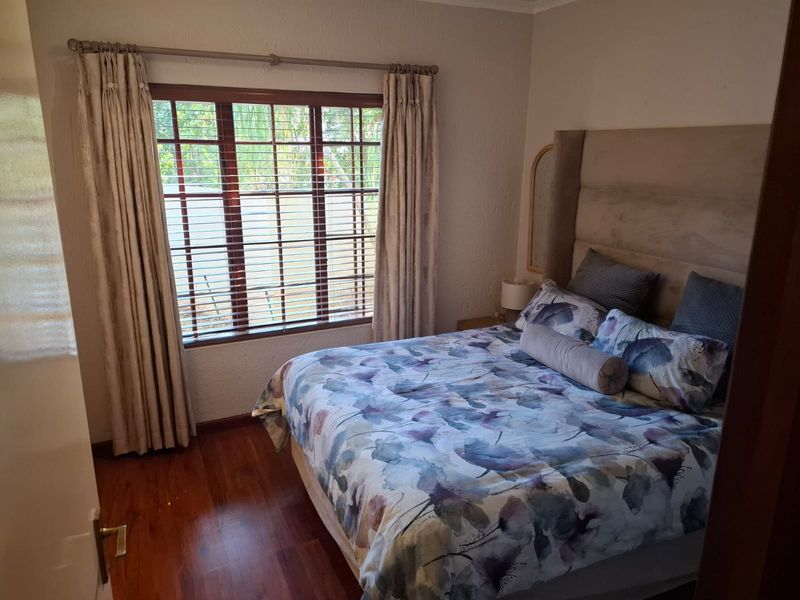 A cozy bedroom featuring warm-toned laminated flooring, a large wooden-framed window with venetian blinds, and neutral wall finishes.