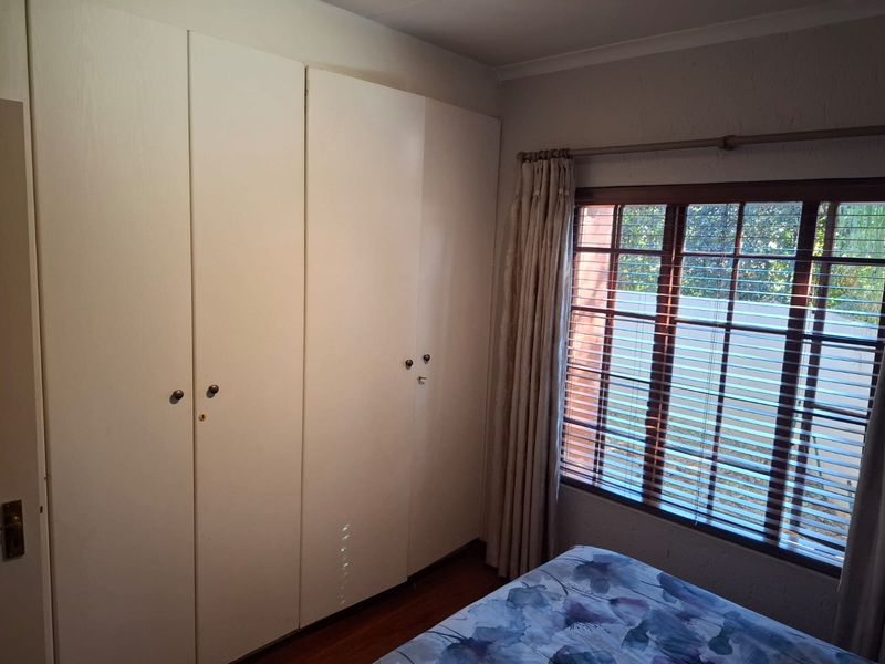 A bedroom featuring built-in cupboards, laminate flooring, and a window with blinds, offering a functional and well-lit living space.