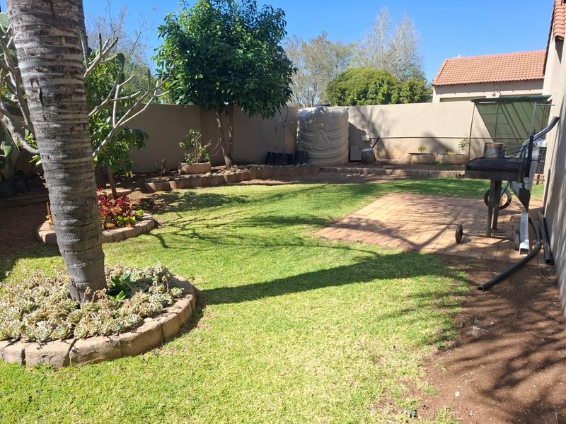 A private rear garden featuring a paved patio area, established trees, landscaped garden beds, and a water storage tank enclosed by boundary walls.