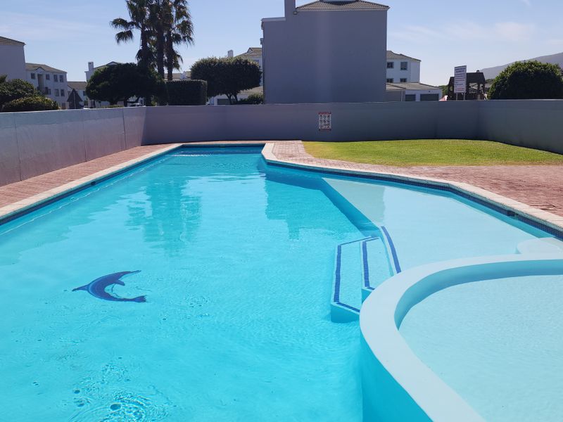 A well-maintained communal swimming pool area featuring paved decking, a boundary wall, and integrated steps, set within a residential apartment complex.