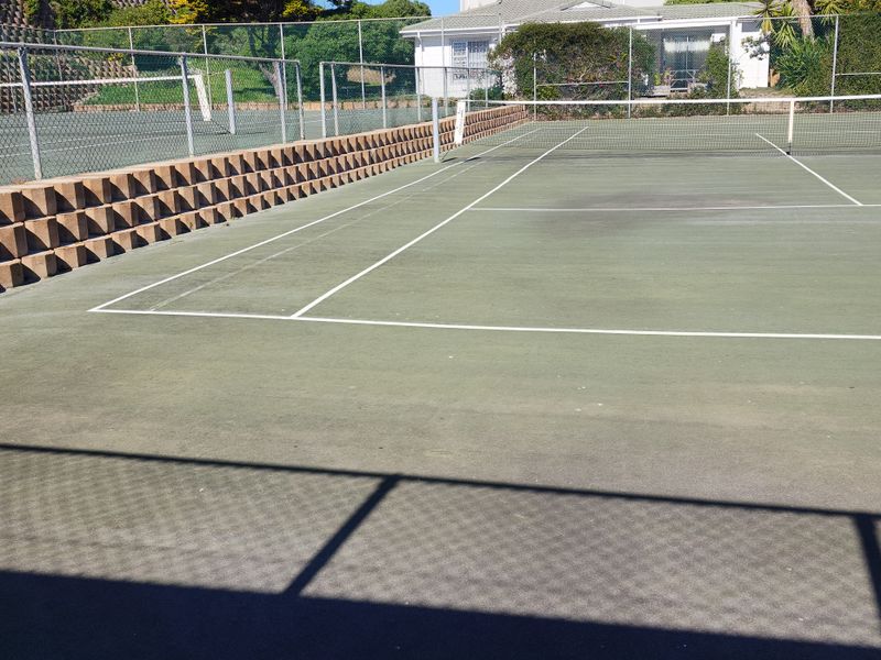A communal tennis court featuring a hard surface, perimeter chain link fencing, and a retaining wall structure.