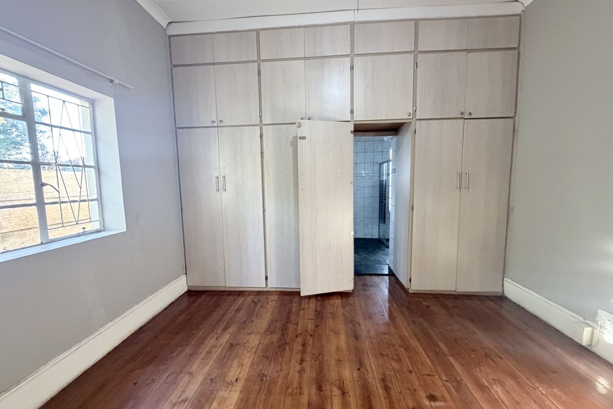 A spacious bedroom featuring polished hardwood flooring, extensive built-in cabinetry, and a window with security bars, leading into an adjoining bathroom.