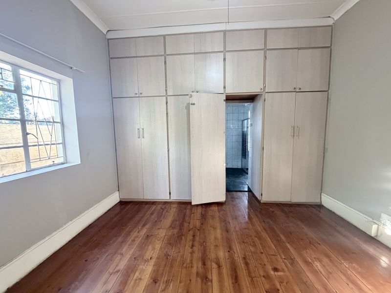 A spacious bedroom featuring polished hardwood flooring, extensive built-in cabinetry, and a window with security bars, leading into an adjoining bathroom.
